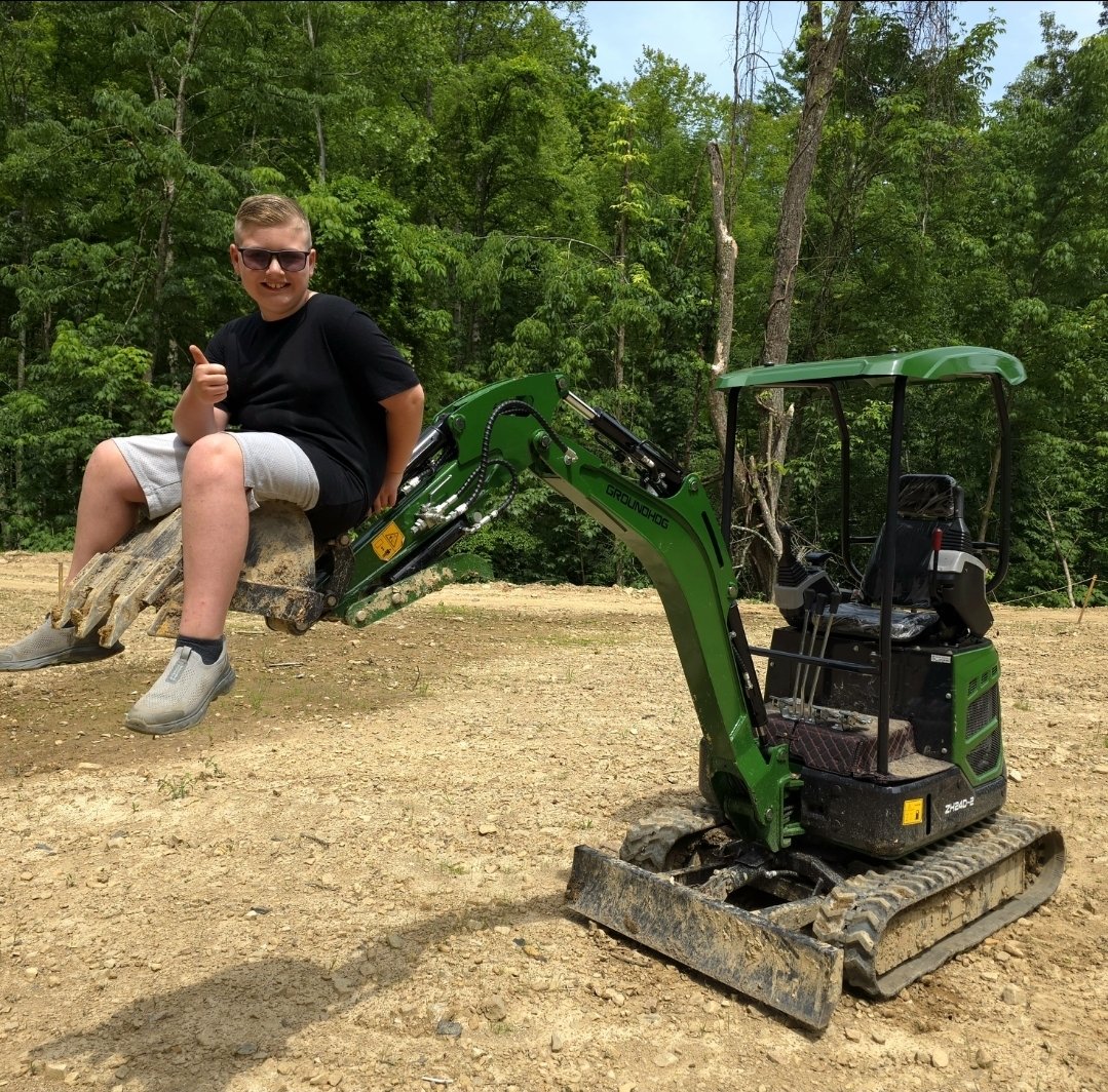 Tayden Franklin sitting in the raised bucket of the green mini excavator, giving a thumbs up